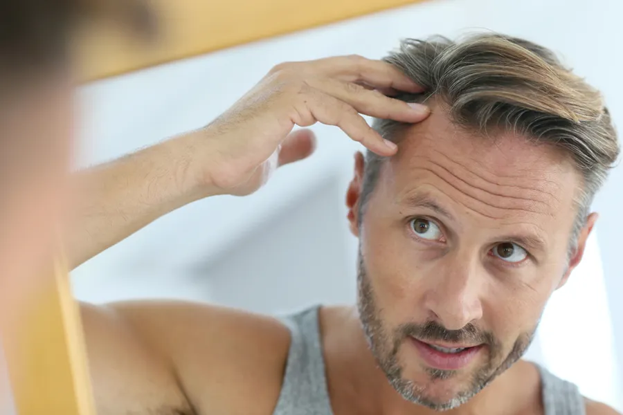 Peptides-for-Hair-Restoration-Doctor A middle-aged man with blonde and gray hair inspects his receding hairline in the mirror, considering getting peptides for hair restoration treatments from Healthy Aging Medical Centers of New Jersey in Northern New Jersey.